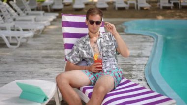 Ağır çekim. Genç bir adam plaj şezlongunda yarı oturarak bardakta bir kokteyl ile poz veriyor, yaz günü havuz kenarında dışarıda. Adam kameraya güneş gözlüklerinden bakıp gülümsüyor.