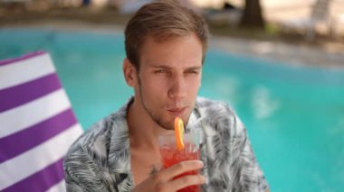 Ağır çekim. Yakın plan. Mutlu genç adam, havuz kenarında bir plaj şezlongunda oturuyor bardaktan kokteyl pipetini içiyor ve gülümseyerek kameraya bakıyor.