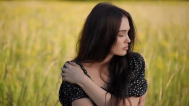 Siyah elbiseli, uzun saçlı esmer bir kadın, açık havada güneşli bir yaz gününde, sarı çayır çimlerinin arka planında durarak kollarıyla kendine sarılıyor. Hoş bir kız.