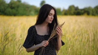 Esmer bir kız elinde bir buket sarı dikenli çubukla poz veriyor, açık havada güneşli bir yaz gününde arka planda sarı çayır çimlerinin önünde duruyor. Siyah elbiseli güzel bir kız.