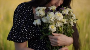 Yakın plan. Koyu renk uzun saçlı genç bir kadın geniş bir buket kır çiçeğini sıkıca kavrıyor ve bir yaz günü açık havada sarı yeşil çayır çimlerine doğru sallanıyor.