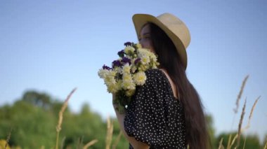 Uzun saçlı, kenarlı bir şapka takan ve elinde bir buket kır çiçeği olan genç bir kadın kameraya doğru dönüyor. Gülümseyen kız, açık havada bir yaz günü tarlada dikilirken kameraya bakıyor.