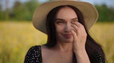 Yakın plan. Şapkalı bir kız elinde kır çiçeğiyle poz veriyor ve gülümseyen kameraya bakıyor. Bir kız, yaz günü sarı bir tarlanın ve yeşil ağaçların arkasında duruyor.