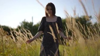 Düşük açılı çekim. Siyah elbiseli, uzun saçlı, tatlı esmer bir kız güneşli bir yaz gününde uzun, sallanan çimlerle bir tarlada yürüyor ve etrafına bakıyor.