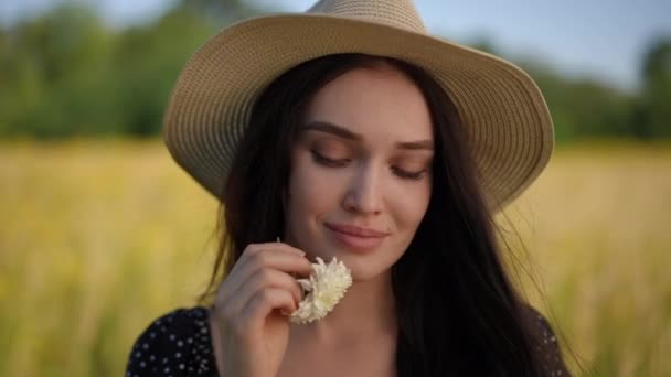 漂亮的年轻女子戴着帽檐 手里拿着野花 微笑着看着相机 夏日里 一个女孩站在一片黄色的田野和绿树的背景下 — 图库视频影像