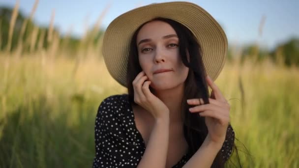 一个戴着帽檐 一头乌黑的长发的漂亮姑娘坐在田野的草地上 带着调情的神气看着摄像机 面带微笑 — 图库视频影像