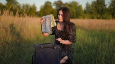 Siyah elbiseli bir kız bavulundan katlanmış kağıt bir harita çıkarıyor. Kız tarlanın ortasına çömelmiş ve nerede olduğunu belirlemek için bir kağıt harita açmış.