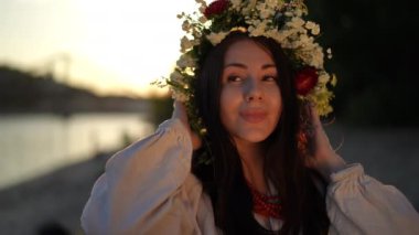 Slow motion. A woman adjusts a wreath of wild herbs and flowers on her head while standing in a traditional embroidered shirt at sunset on a summer evening on the river bank. The woman looks away and