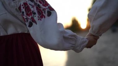 Slow motion. Rear view. Close-up. Two unrecognizable women walk hand in hand on a sandy beach at sunset. The women are wearing traditional embroidered shirts and red Ukrainian ethnic skirts