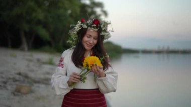 A woman in an ethnic traditional embroidered dress and a wreath of wild flowers on her head inhales the aroma of a bouquet of yellow-orange chrysanthemums in her hands while walking along the river