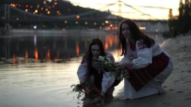 Feast of Ivan Kupala on the Dnipro River. Women in traditional Ukrainian costumes squat and place wreaths of wild flowers and herbs in the river water on a summer evening against the backdrop of the