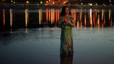 A mysterious girl in a green dress stands in the river water, holds a burning candle in a candlestick in her outstretched hand and sings quietly. A girl poses on a night against the background of the