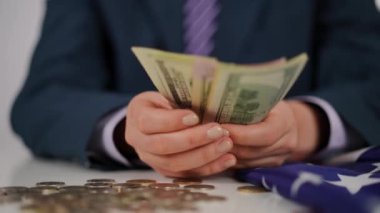 Yakın plan. Mavi takım elbiseli, gömlekli ve kravatlı tanınmayan bir adam Amerikan dolarlarını elinde tutuyor ve dağınık nikel ve bakırdan oluşan bir arka planda beyaz bir masada oturuyor.