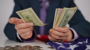 Yakın plan. Mavi takım elbiseli tanınmayan bir insan, beyaz bir masada, dağınık nikel ve bakır paraların ve katlanmış Amerikan bayrağının önünde otururken Amerikan dolarlarını sayıyor.