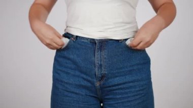 Orta boy yakın çekim. Beyaz tişörtlü ve kot pantolonlu tanınmayan bir kadın ceplerini boşaltıyor, kameraya boş olduklarını gösteriyor, beyaz bir arka plana karşı içeride dururken