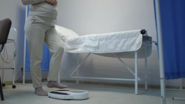 Pantolon ve çorap giymiş tanınmayan hamile bir kadın klinikte yerde duran banyo terazisinde duruyor. Hamilelik sırasında tartılıyor. Hamilelik ağırlık kontrolü