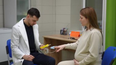 Beyaz önlüklü bir erkek doktor, acil serviste karşısında oturan hamile bir kadının parmağına nabız oksimetresi sensörü takıyor. Doktor hastaları ölçer.