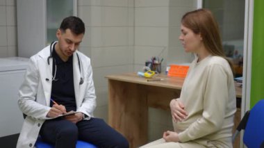 Bir doktor, ambulans çağırdıktan sonra hamile bir kadının ifadesini not alıyor. Erkek bir doktor dikkatle dinler, gülümser ve not defterine bir kalemle yazar.