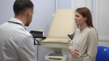 Ağır çekim. Ambulans odasındaki jinekoloji sandalyesinin yanında oturan bir doktor ve hamile bir kadın konuşuyor.