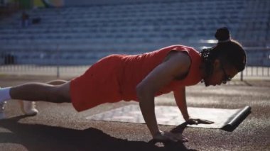 Orta boy yakın çekim. Kırmızı spor giyimli, güneş gözlüklü ve rastalı genç bir sporcunun profili, bir sonbahar günü stadyumda koşu pistinde spor yaparken, fitness platformu 9 kullanarak şınav çekiyor.