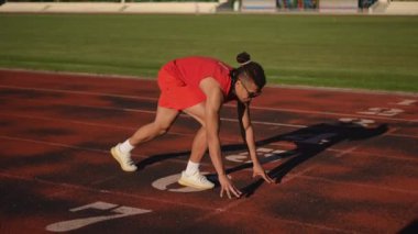 Kırmızı spor giyimli, güneş gözlüklü ve rastalı bir erkek sporcu çömelme pozisyonunu alır ve öne doğru koşmaya başlar..