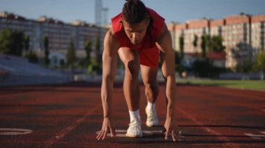 Ağır çekim. Yakın plan. Kırmızı spor giyim ve rasta giymiş bir erkek sporcu bir stadyumun koşu yoluna çömelmiş ve kameraya bakıyor. Kamera kameraya yakınlaşıyor.