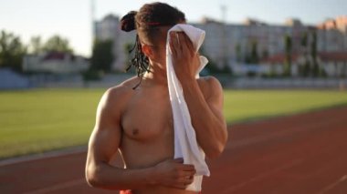 Ağır çekim. Orta boy yakın çekim. Kafasında rasta olan bir sporcu uzun bir idmandan sonra beyaz havluyla yüzünden ter siler ve derin derin nefes alır. Elinde havluyla dışarıda duran atletik bir adam.