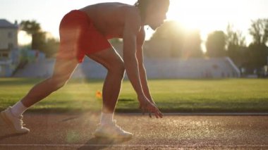 Ağır çekim. Güneşe karşı ateş ediyorlar. Kırmızı erkek atletik şortlu rastalı bir koşucu, güneşli bir sonbahar gününde çömelmiş bir şekilde stadyuma doğru koşar.