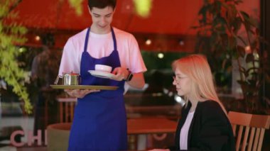 Ağır çekim. Garson bir kız getirdi ve masaya porselen bir bardak ve metal bir çaydanlık koydu. Bir şehir kafesinin terasındaki masada oturan bir kıza. Uzun boylu sarışın kız.