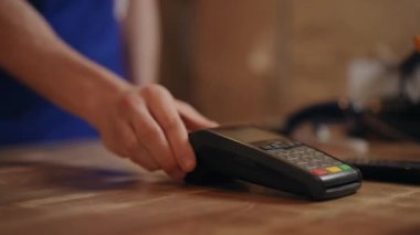 Yakın plan. Tanımlanamayan mavi önlüklü bir adam POS terminalini eliyle itiyor ve tanınmayan bir kadın cep telefonunu üzerine koyup bir ödeme yapıyor.