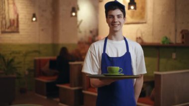 Mavi önlüklü ve mavi beysbol şapkalı bir garson yeşil çay tabağı ve fincanla tepsiyi tutar ve modern bir kafenin içinde gülümseyen kameraya bakar.