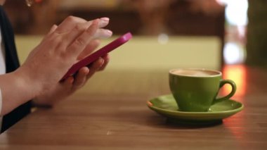 Yakın plan. Tanımlanamayan bir kadın bir kafede bir masada oturuyor ve elinde bir cep telefonu tutuyor, ekranda parmağıyla tıkırdıyor ve kaydırıyor. Kadının biri arka planda cep telefonu tutuyor.