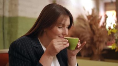 Bir kadın iki eliyle yeşil bir bardak kapuçino tutuyor ve gözlerini kapatıp kahve kokusunu içine çekiyor. Bir kadın kafede bir masada otururken porselen bardaktan kahve içer.