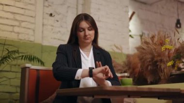 Bir iş kadını, bilek akıllı saat ve iç çeker, bir kafede bir masada otururken, başını tutarak gergin bir şekilde bakar.