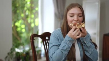 Kot ceketli bir kız yemek masasındaki ahşap bir sandalyeye oturur ve elleriyle tuttuğu pizzayı yer. Genç kız pizzayı seviyor, gülümsüyor, kameraya başparmağıyla bakıyor.