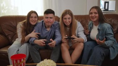 Bir grup arkadaş, oturma odasındaki deri koltukta oturup konsolda oynarken eğleniyorlar. Elinde oyun tahtası olan bir kız yanında oturan adama karşı bir video oyunu kazanıyor.