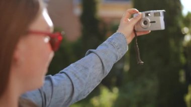 Omzunun üstünden. Güneş gözlüklü bir kız, güneşli bir yaz gününde dışarıda beklerken eski bir fotoğraf makinesiyle selfie çekiyor..