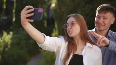 Güneş gözlüklü bir kız, güneşli bir yaz gününde dışarıda dikilirken cep telefonu kamerasıyla erkek arkadaşıyla selfie çeker. Poz veren, gülümseyen ve elleriyle el kol hareketi yapan bir çift genç.