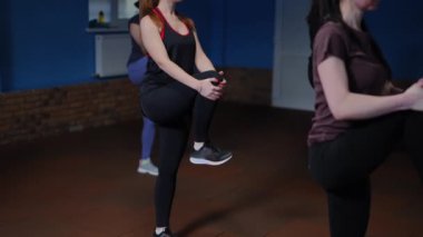 Ağır çekim. Spor giyimli bir grup kadın yavaşça ayaklarını çeviriyor, bir dizlerini göğüs hizasına kaldırıyor ve iki eliyle tutuyor. Kadınlar spor salonunda içeride dururken spor yapıyor.