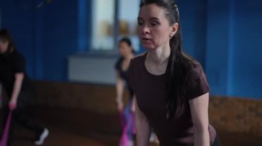 Bir kadın antrenör spor giyimli bir grup kadına komuta ediyor, tek dizle çömelme hareketi yaparken, elastik bir fitness direnç bandı kullanıyor. Kadınlar spor salonunda grup antrenmanı yapıyor.