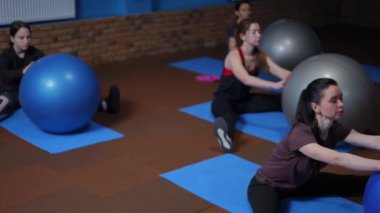 Ağır çekim. Spor giyimli bir grup kadın spor kıyafetleri içinde bacakları açık bir şekilde fitness minderlerine oturur ve elleriyle bir fitball topunu hareket ettirirler. Jimnastik topu kullanarak spor yapmak.