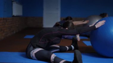 Ağır çekim. Spor giyimli kadınlar, geniş bacaklı fitness paspaslarının üzerinde oturuyorlar, spor toplarını elleriyle hareket ettiriyorlar ve öne doğru eğiliyorlar. Jimnastik topu kullanarak spor yapmak.