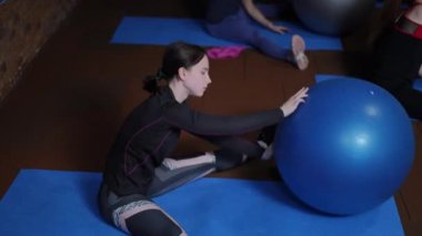 Spor giyimli kadınlar, geniş bacakları olan fitness paspaspaslarının üzerinde oturuyorlar, yanal viraj yapıyorlar ve elleriyle spor toplarını oynatıyorlar. Jimnastik topu kullanarak spor yapmak.