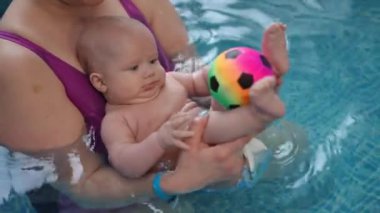 Yakın plan. Annesinin kollarındaki bir bebek yüzme havuzunda suda otururken çok renkli küçük bir futbol topuyla oynuyor.