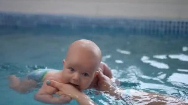 Yakın plan. Bebek, annesine yüzerken, çocuk havuzunda kollarını açmış kameraya bakıyor.