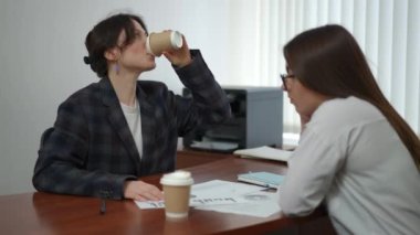 Spor ceketli bir iş kadını tek kullanımlık plastik bardaktan kahve içer ve masasında uzanan bir programı bir bayan meslektaşıyla tartışır. Kadınlar ofiste bir masada otururlar.