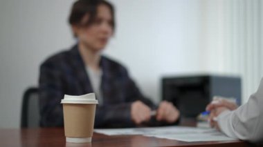 Ön planda, ofisteki bir masada kahve standı olan tek kullanımlık bir kağıt bardak. Arka planda zayıf bir defocus, spor ceketli bir kadın yönetici bir kağıt bardaktan kahve içer ve bir