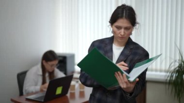 Ön planda, spor ceketli bir kadın elinde plastik bir klasör tutuyor ve ofiste dururken içindeki kağıt belgelere bakıyor. Arka planda, bir kız masasına oturur ve kitap okur.