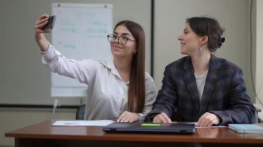 Gözlük takan bir kız, içeride bir masada otururken kız arkadaşıyla cep telefonu kamerasıyla selfie çeker. Kızlar cep telefonu kamerasına bakıp gülümsüyor