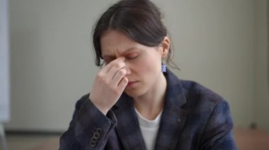 Yakın plan. Spor ceketli bir kadın gözlerini kapatır, parmaklarıyla burun köprüsünü kavrar, ellerini tutar, ofiste otururken baş ağrısını hafifletmeye çalışır..